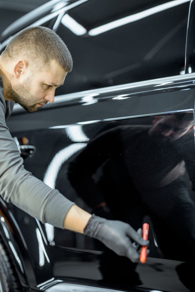 Worker examining vehicle body for scratches and damages backliting by a flashlight at the service. Checking car paint before vehicle detailing